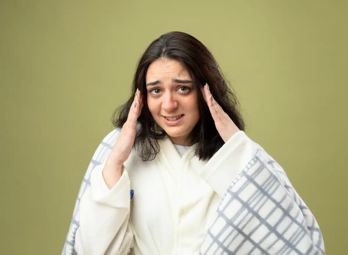 A young woman wrapped in a blanket holds her temples with a worried expression, suggesting headache, stress, or feeling unwell.