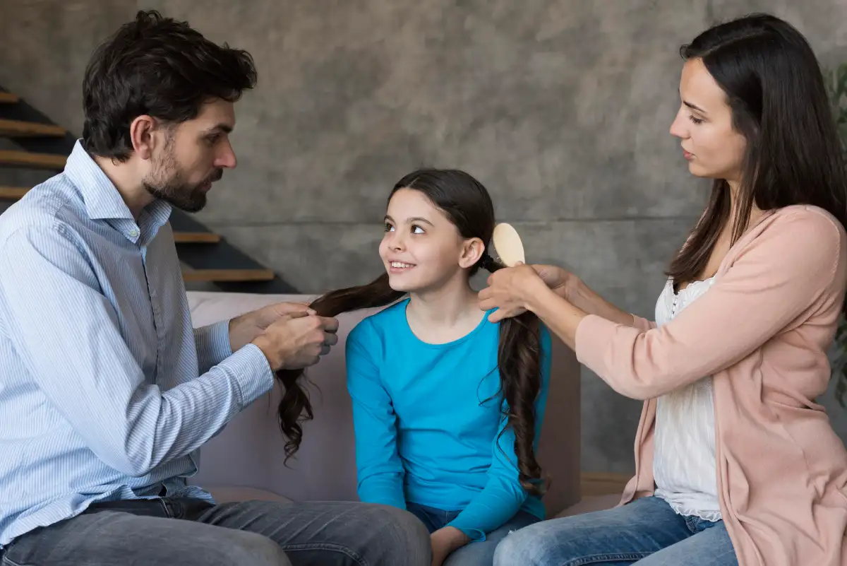 Parents helping their young daughter get ready, with father holding her hair and mother brushing it while the child smiles at home