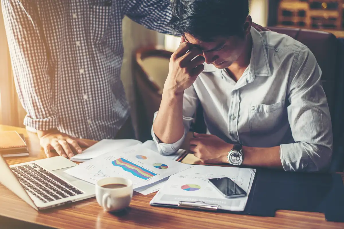 Stressed businessman reviewing financial reports at a desk with laptop, charts, and coffee while a colleague stands nearby offering support