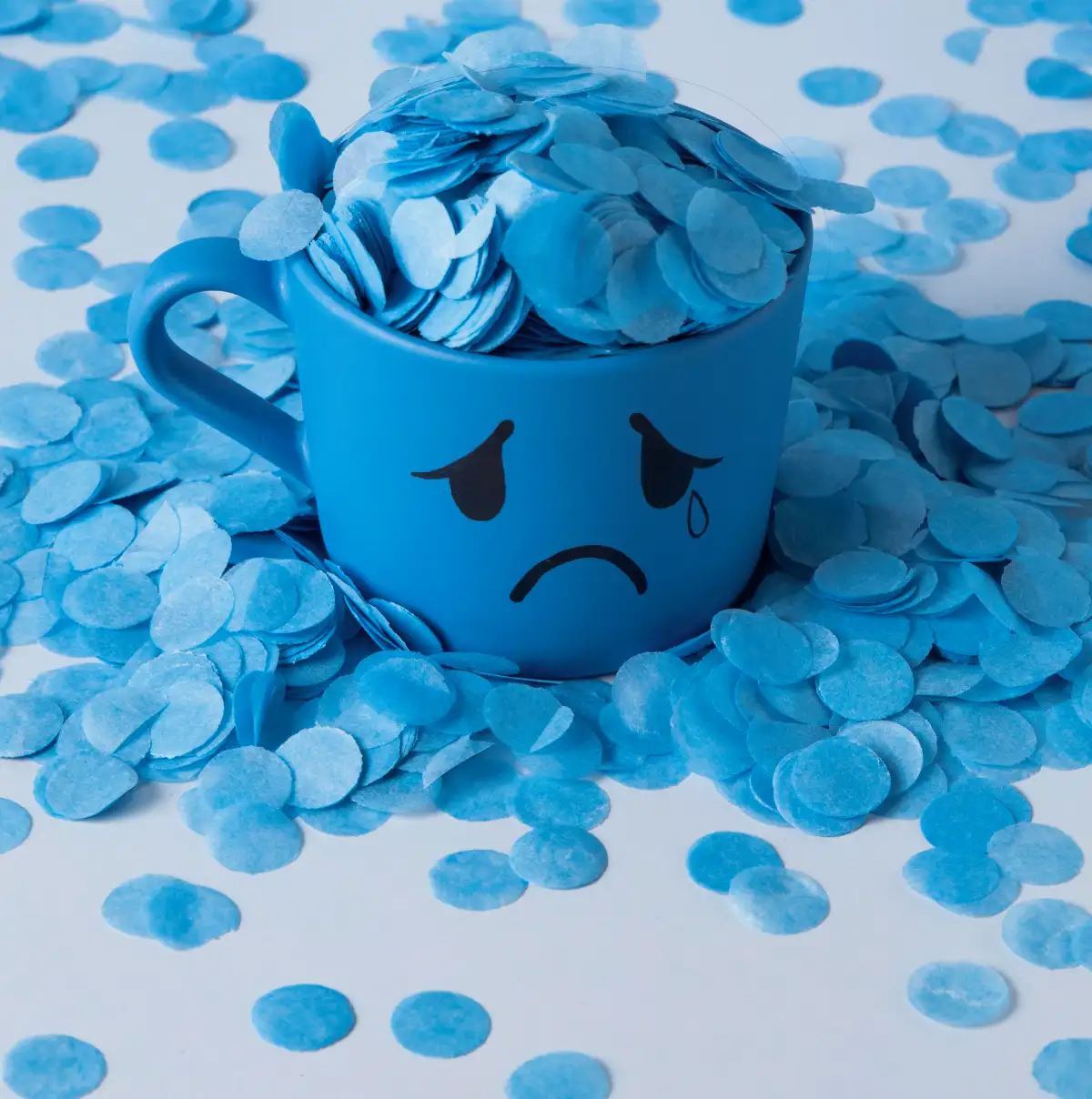 Blue cup with a sad crying face overflowing with blue paper confetti, symbolising stress, sadness, anxiety, and emotional overwhelm.