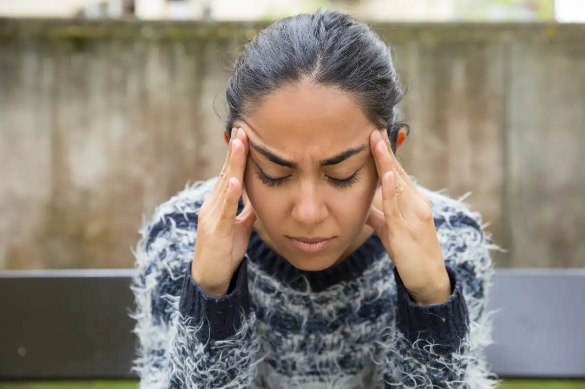 A stressed young woman sitting on a bench outdoors, holding her temples with a worried expression, appearing overwhelmed or anxious.