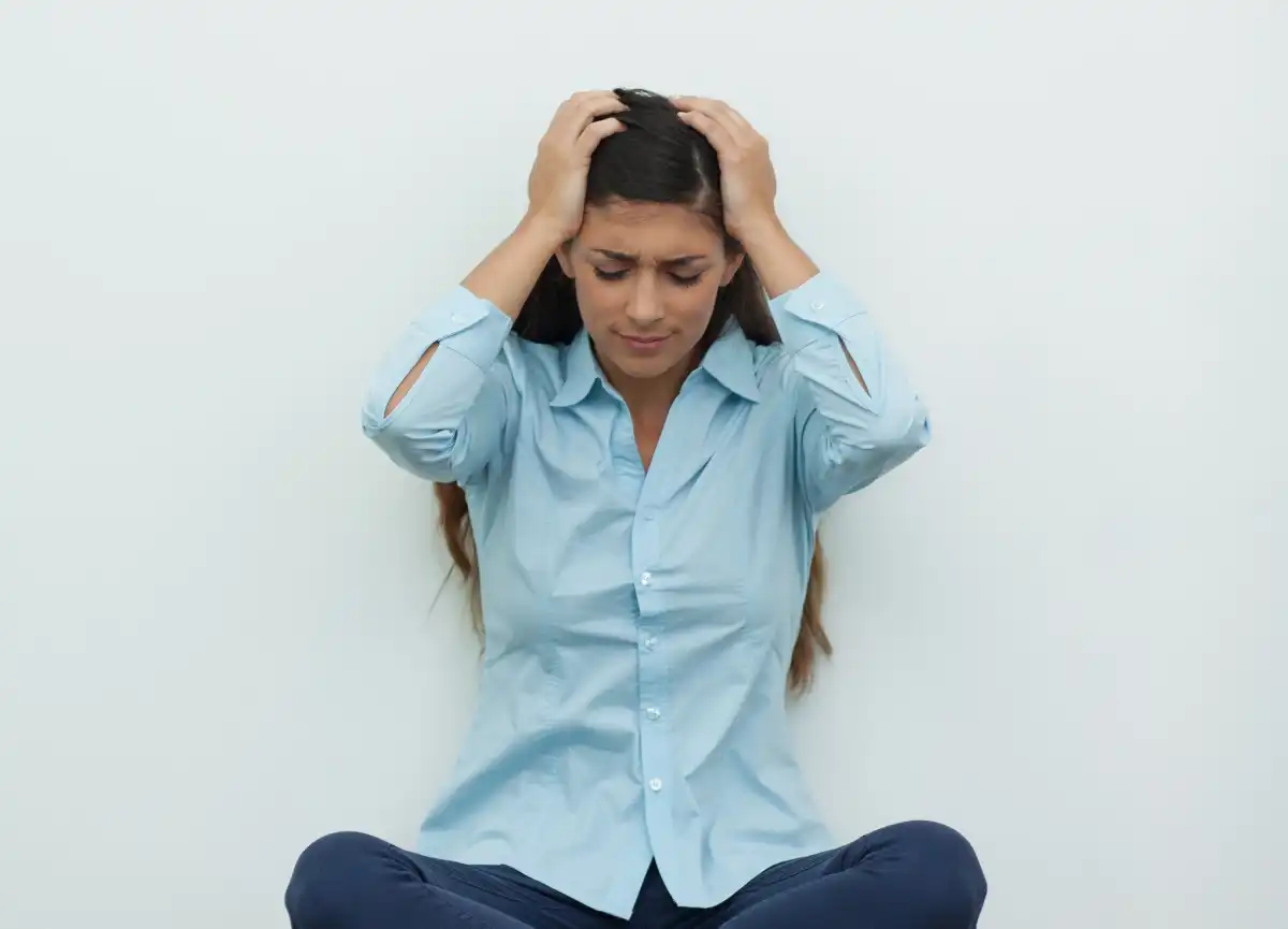 A woman sits cross-legged on the floor against a plain gray wall, holding her head with both hands and looking down, appearing stressed or overwhelmed.