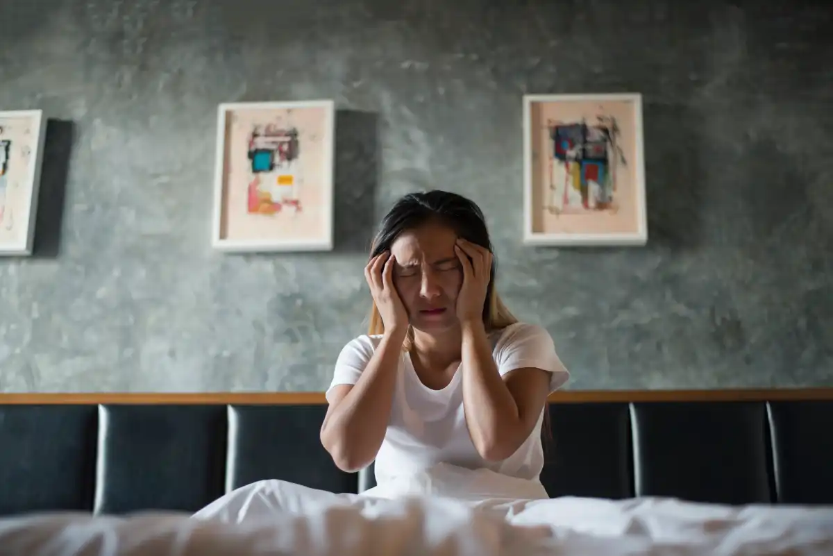 Young woman sitting on a bed with her hands on her face, appearing stressed, anxious, or emotionally overwhelmed indoors.